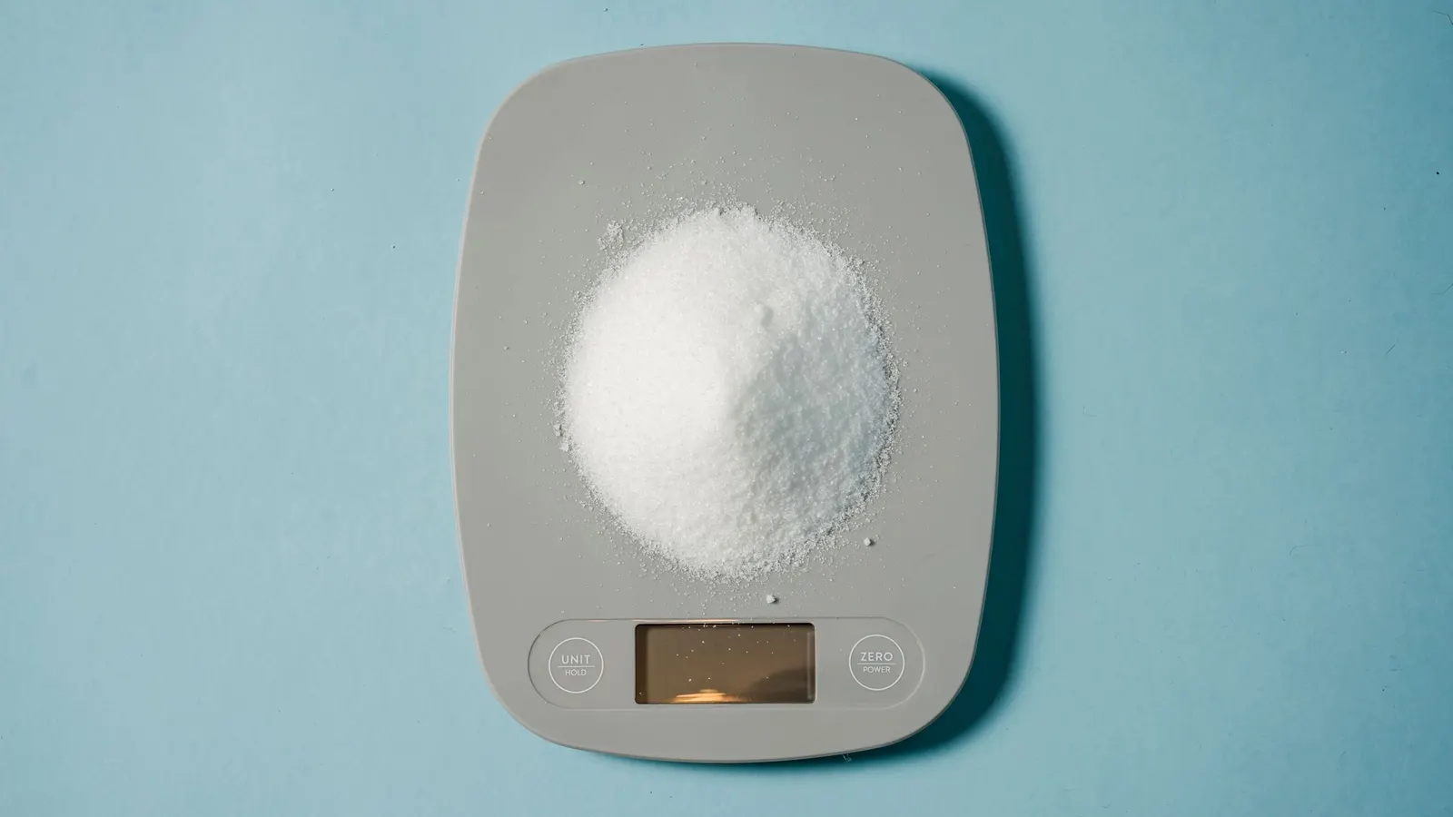 Mound of white granulated sugar on a gray digital kitchen scale against a pale blue background