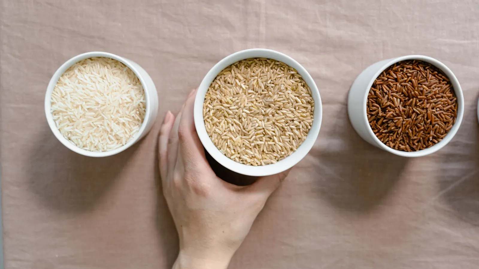 Hand picking up a bowl of brown rice between bowls of white rice and red rice