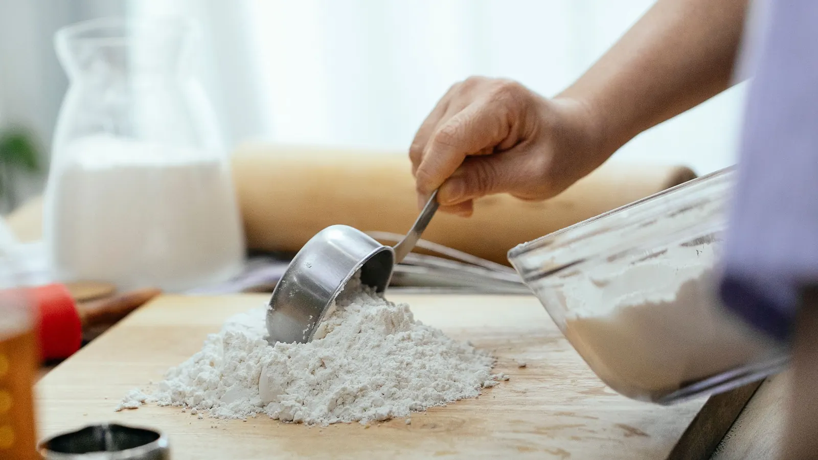 Hand using a metal measuring cup to scoop flour onto a wooden board