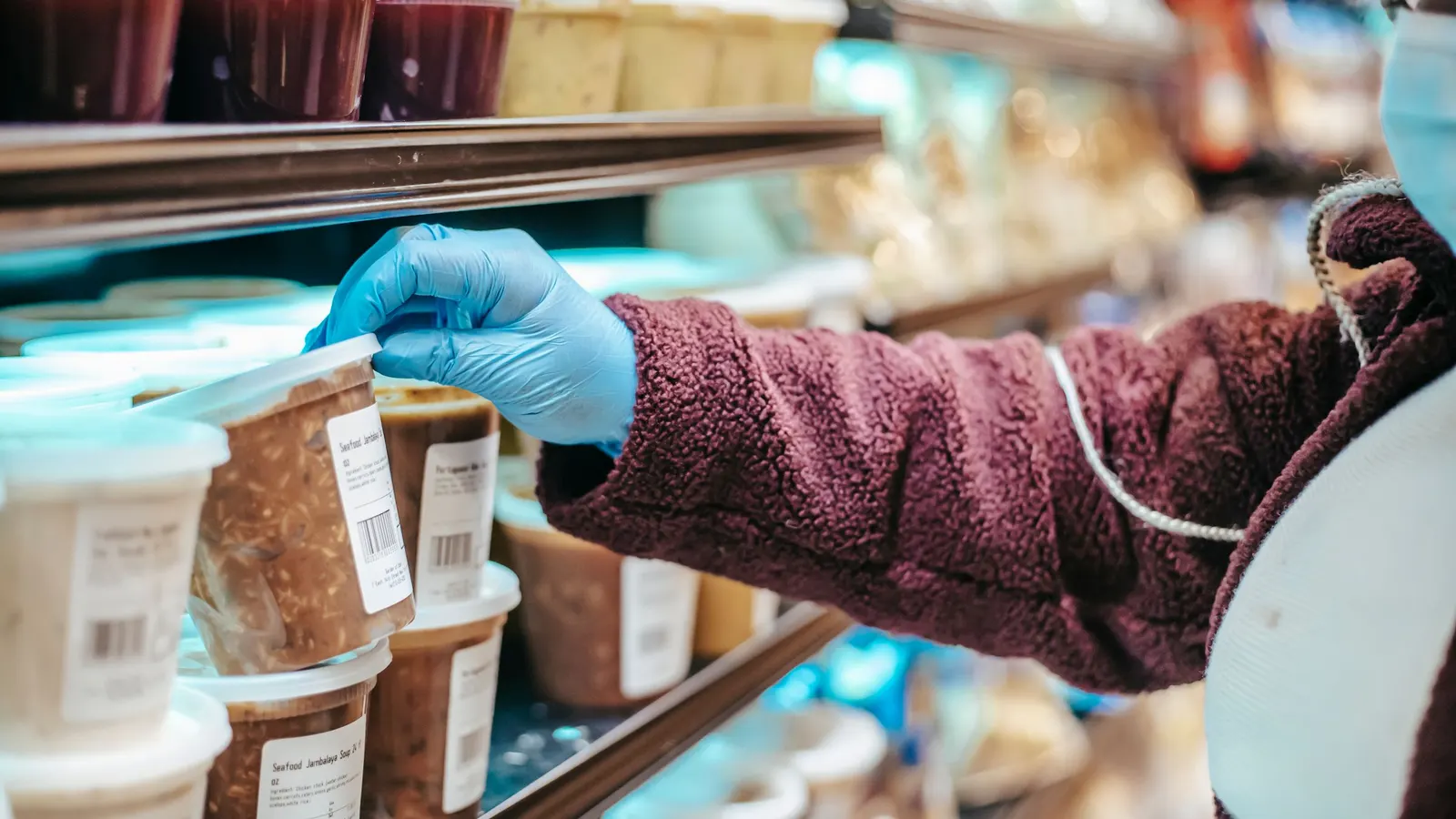 Gloved hand reaching for labeled deli containers stacked on a refrigerated store shelf