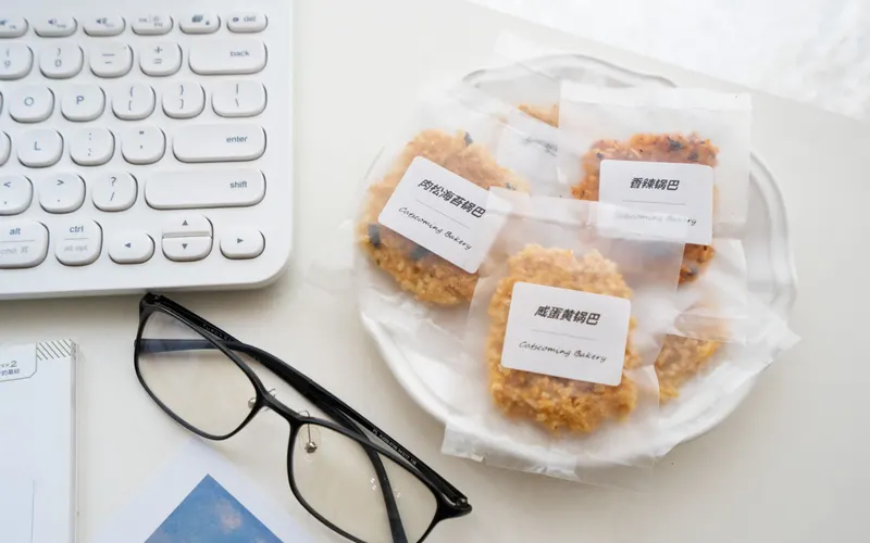 Individually bagged cookies with printed white labels on a plate beside a keyboard and glasses