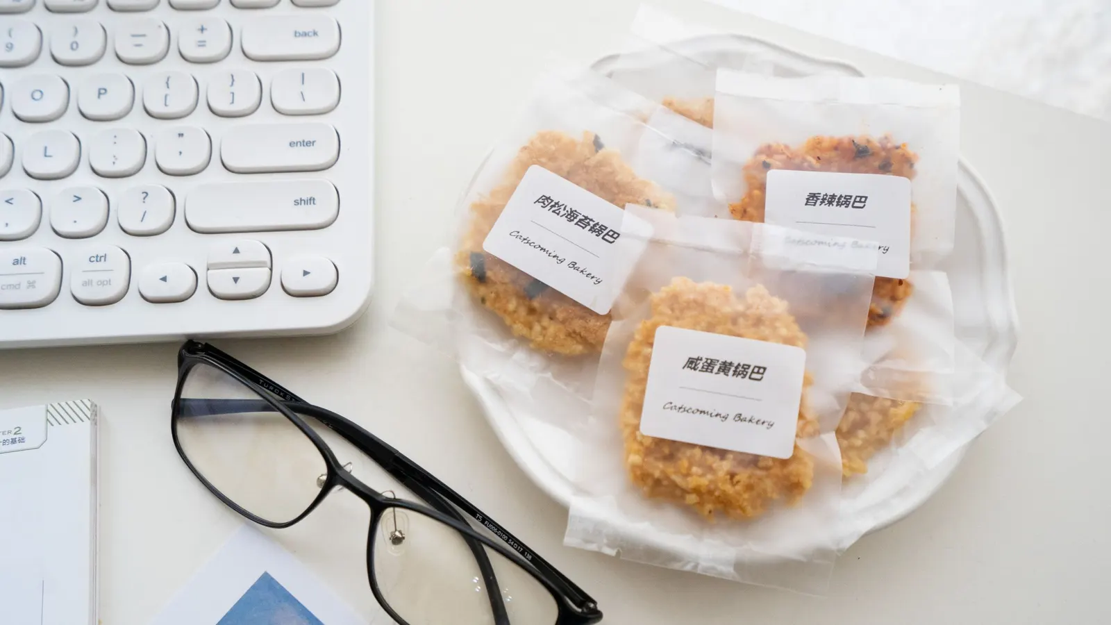 Individually bagged cookies with printed white labels on a plate beside a keyboard and glasses