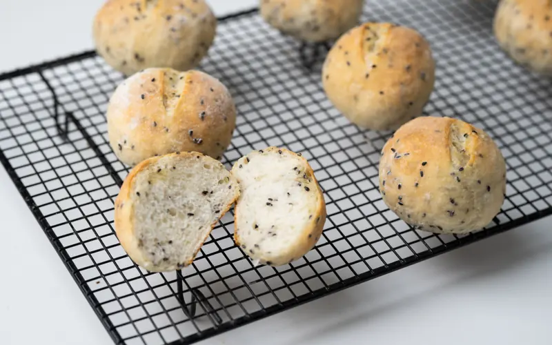 Seeded bread rolls cooling on a wire rack, one broken open to show the crumb
