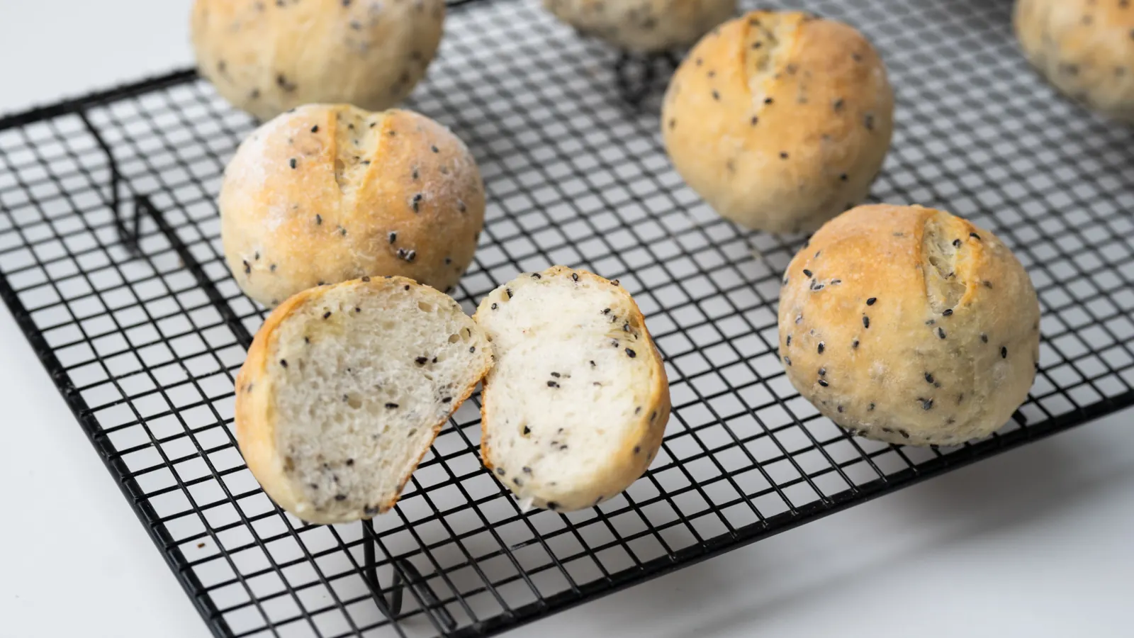 Seeded bread rolls cooling on a wire rack, one broken open to show the crumb