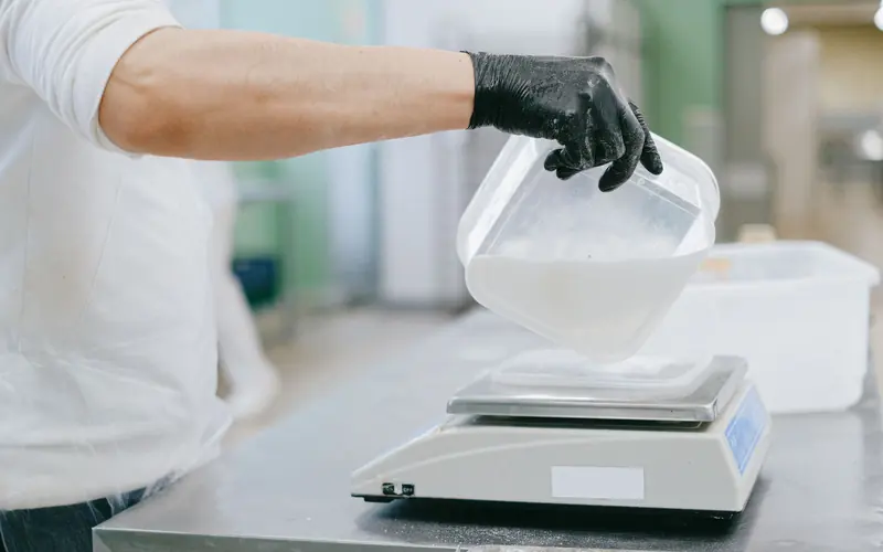Gloved hand pouring a dry ingredient into a container on a digital scale in a production kitchen