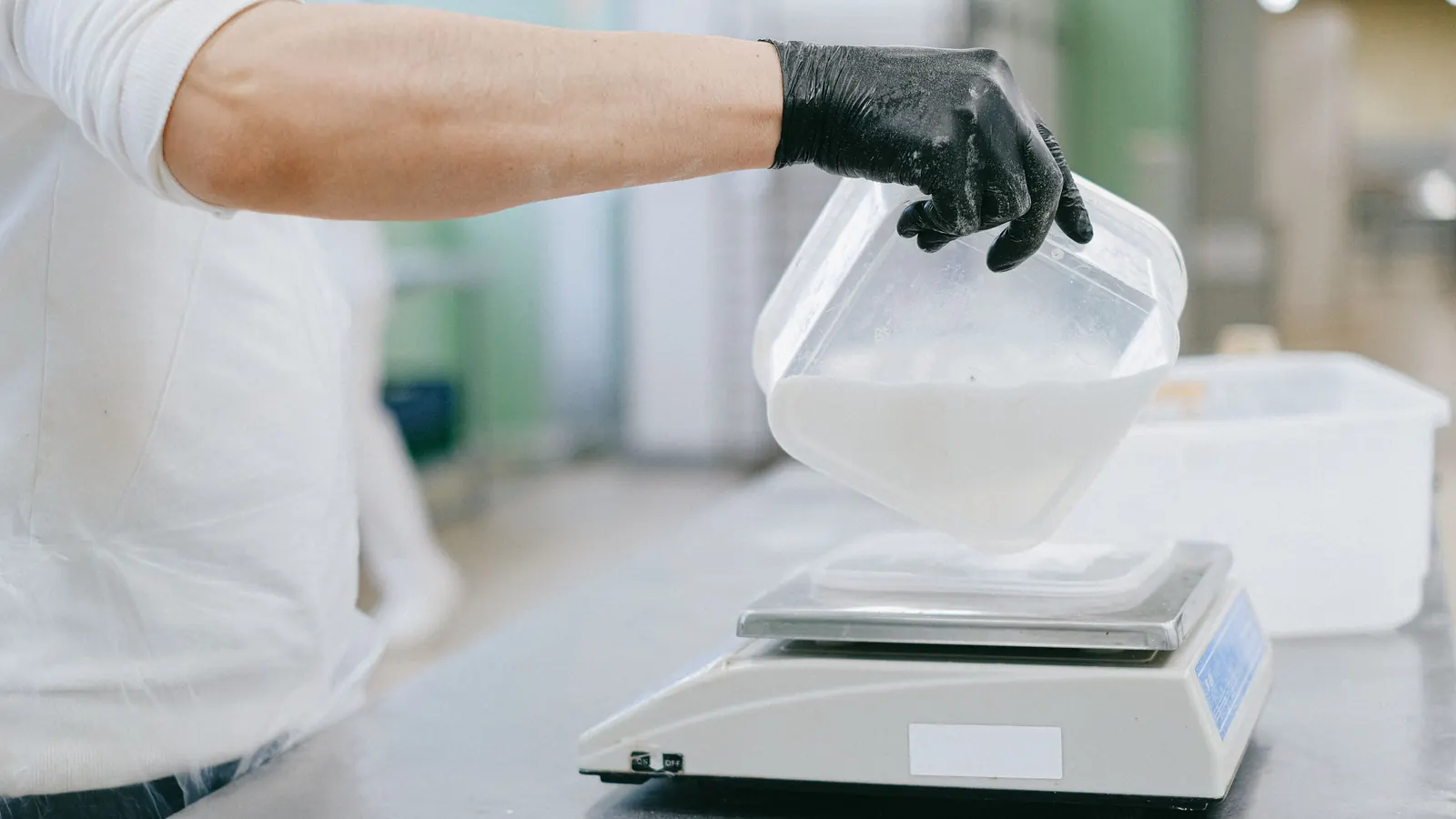 Gloved hand pouring a dry ingredient into a container on a digital scale in a production kitchen
