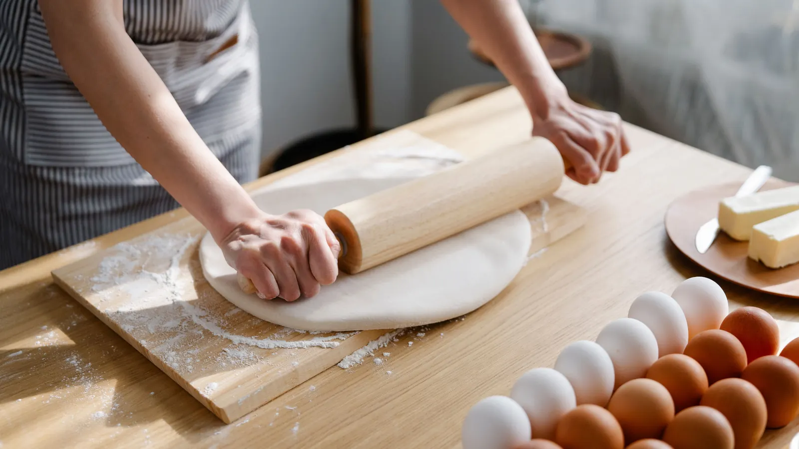 Hands rolling out dough on a wooden board beside a tray of eggs and a dish of butter